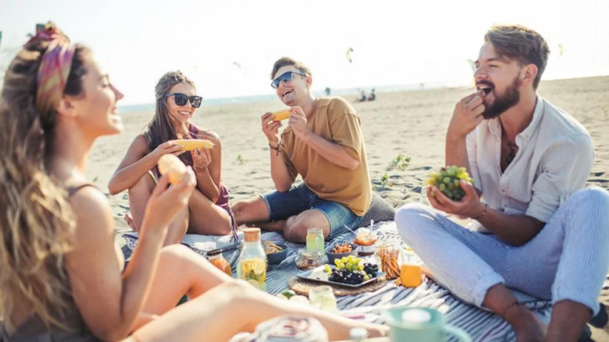 Grupo de amigos comiendo en la playa con recipientes térmicos para comida.